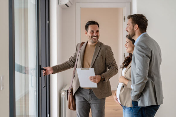 The photo shows a cheerful real estate agent welcoming a couple into a home. The agent, standing near a large glass door, is smiling while holding the door open with one hand and a clipboard in the other. He wears a brown blazer, beige sweater, and carries a shoulder bag. The couple, dressed smartly, stand nearby and smile as they look toward the agent. The setting appears bright and modern, suggesting a home tour or property showing in progress.