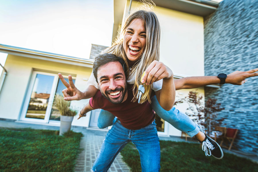 DavideAngelini Happy young couple holding home keys after buying real estate - Husband and wife standing outside in front of their new house