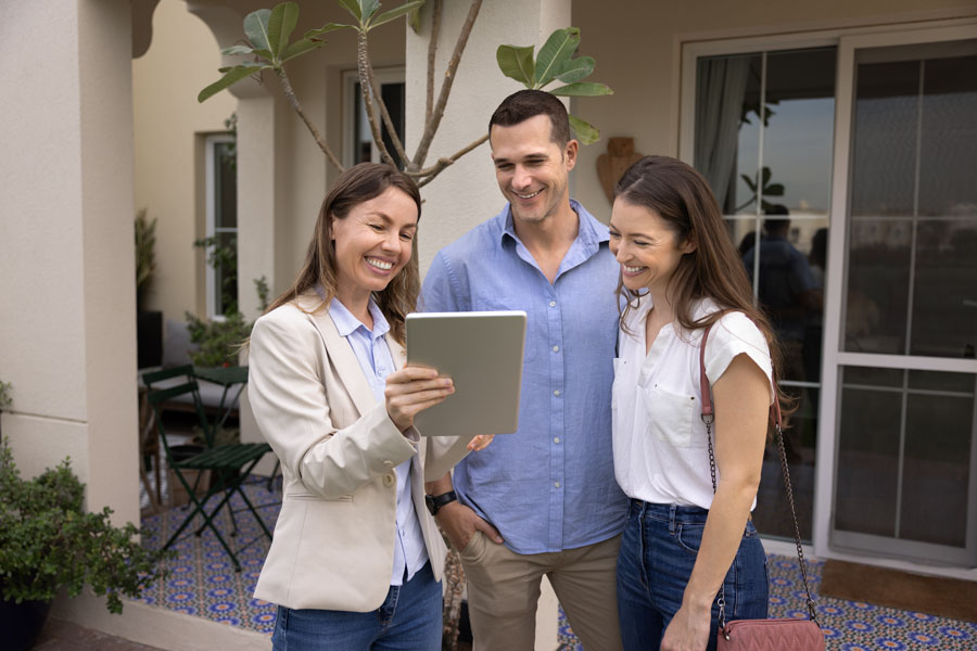 Happy real estate agent woman showing content on tablet to young couple of tenants, renters, house buyers, standing at apartment entrance outside, talking, laughing, discussing rent or purchase terms