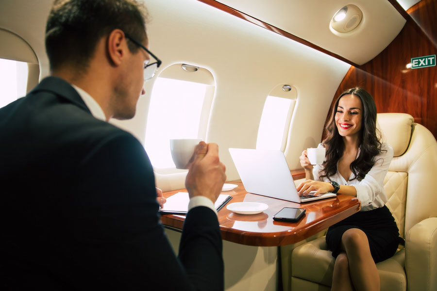 Movers and shakers. A lovely woman in formal attire is typing something on her laptop and drinking coffee while sitting near her male colleague in a window seat in a business class.