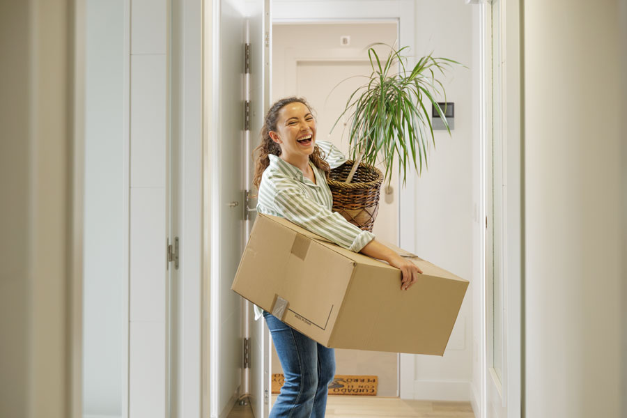 Happy Single pretty young adult woman arriving entering the door of her new moving house with boxes and a plant, real estate, house selling